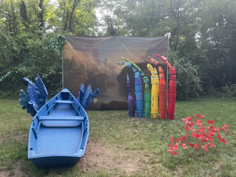 In a wooded area, a theatrical backdrop is set up with a tableau in front of it of a bright-colored, faux rainbow on the right and a bright blue rowboat and faux waves aiming at the picture as if ready to shoot the rapids in the river on the left. Fluttering red butterflies swirl in the foreground. 