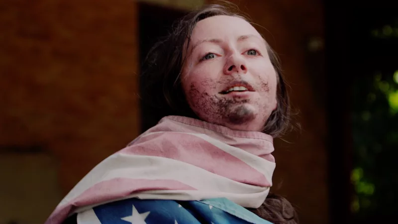An image from Alex Tatarsky's Gnome Core in which she has a faded American flag wrapped around her neck and dirt covering her face.