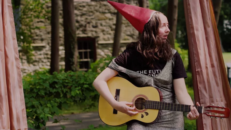 Alex Tatarsky performing Gnome Core, wearing a t-shirt under a silver sparkly dress and a pointed red cap playing guitar in a garden landscape.