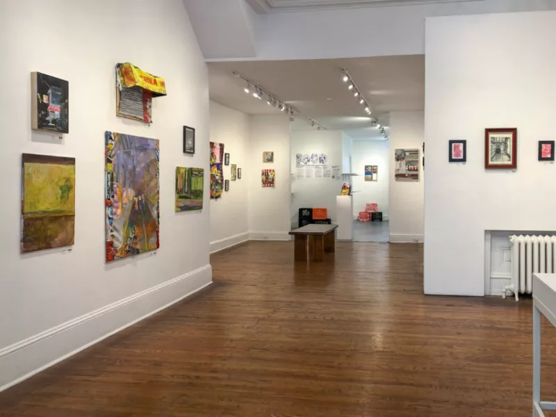 Image of the gallery space at Da Vinci Art Alliance showing Corner Stories an exhibition and food drive by Eustace Mamba, Jake Weiss, and Nasir Young. On the left wall a grouping of five works is arranged, as we go deeper into the space more paintings and drawings appear as well as a bench and podium with sculptural object atop.