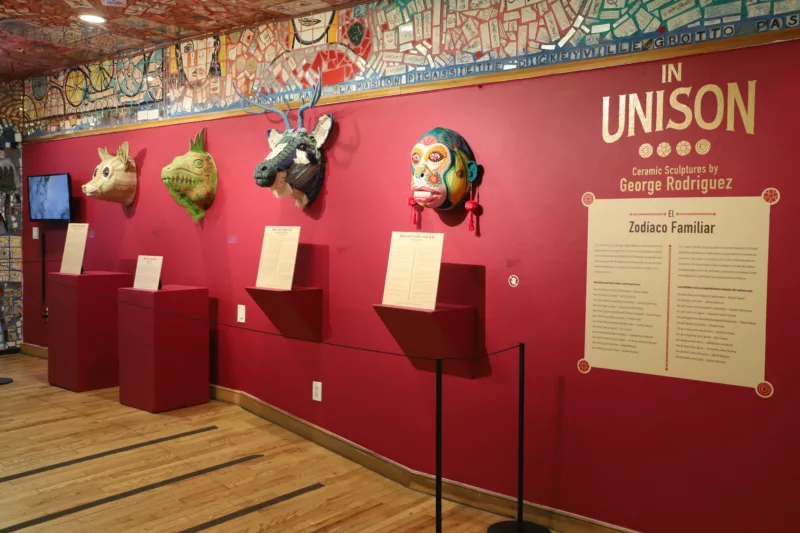 A red gallery wall of Philadelphia's Magic Gardens with mosaic over the top edge of the wall and the entire ceiling. Animal heads line the wall. Covered in patte4rns and decoration expressing individuality and culture.