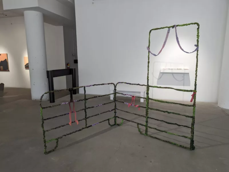 An animal pen covered over by moss ad pink and purple laces in a gallery space with other art works in the background along the white walls.