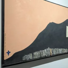 A black mountain with a foreground of rocks creating a smaller mountain. The horizon is a soft peachy color. A note is tacked onto the black mountain. To the left of the canvas is a blue Fleur De Lis and the Artists Signature.