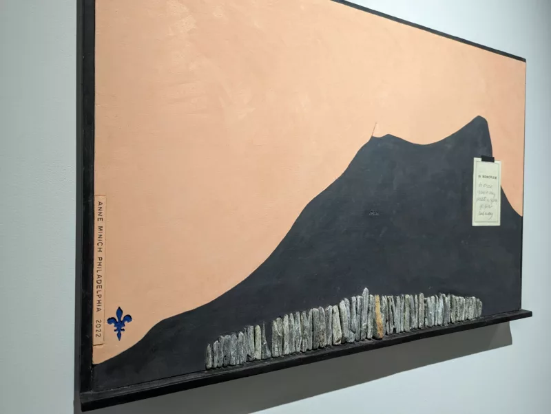 A black mountain with a foreground of rocks creating a smaller mountain. The horizon is a soft peachy color. A note is tacked onto the black mountain. To the left of the canvas is a blue Fleur De Lis and the Artists Signature.
