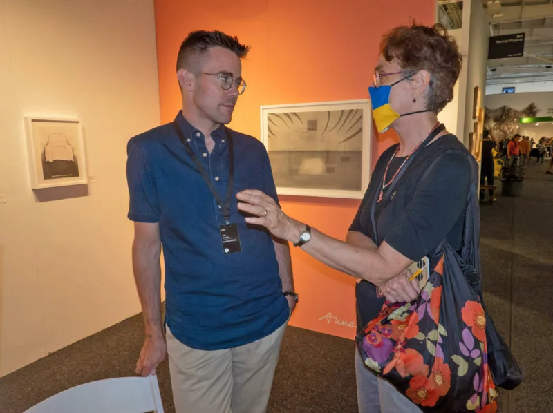 A man in a dark blue, button-down shirt listens while a woman wearing a mask speaks. They are in a booth by Commonweal Gallery showing drawings by Anne Minich on the walls painted orange and cream.