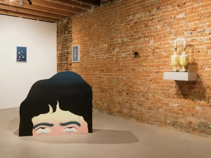 A gallery with a cement floor, exposed brick walls and exposed beams in the ceiling shows a group of works including a large partial cut-out head that seems sunk into or rising out of the floor. On the walls are framed pieces and on a small shelf are two yellow ceramic vessels side by side that seem connected by a kind of ceramic rainbow of all white between them.