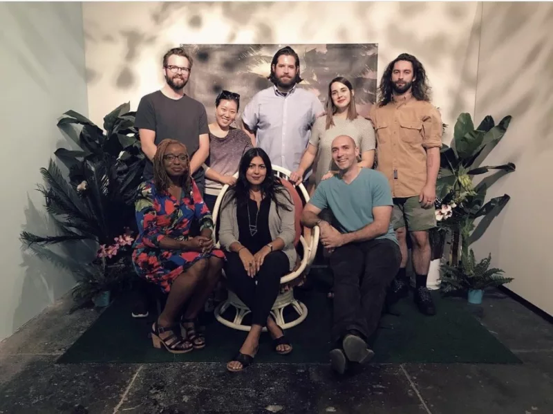 A group photo shows 8 people posing for a picture, smiling for the camera. They sit or stand in a small alcove in a gallery space, Fjord, and they are all members of the artist collective. They look very happy and at ease and the light in the photo has a sparkly glow that elevates the photo beyond a regular group photo into a more artistic realm.