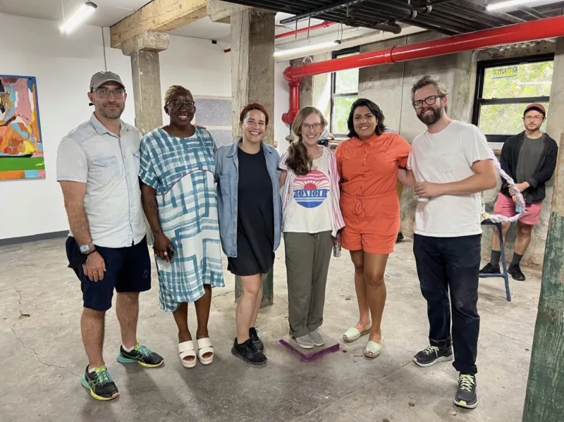 A group portrait photograph of 6 people standing and smiling is a gallery space. They are members of the artist collective, Fjord. In the background is another person, watching. The photo conveys joy and happiness the group, which is diverse in gender and race.