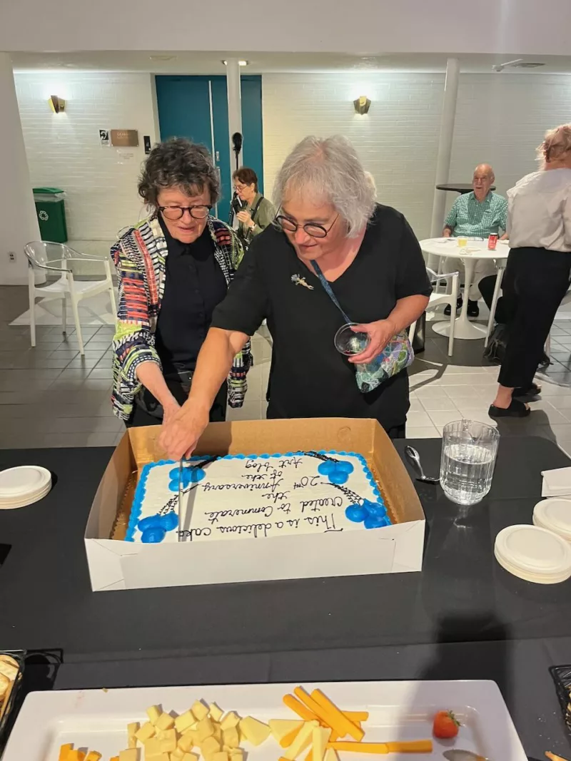 Two women hold a knife and cut cake at a celebration of their 20-year partnership in an arts publication, Artblog.
