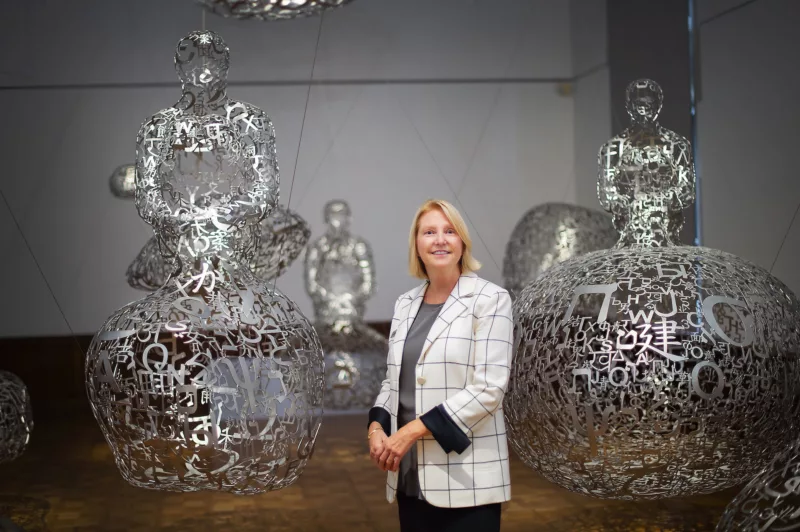 A white woman wearing a white plaid jacket and black pants smiles broadly. She is standing in front of large sculptures in an indoor gallery space. She just retired from duties at the gallery.