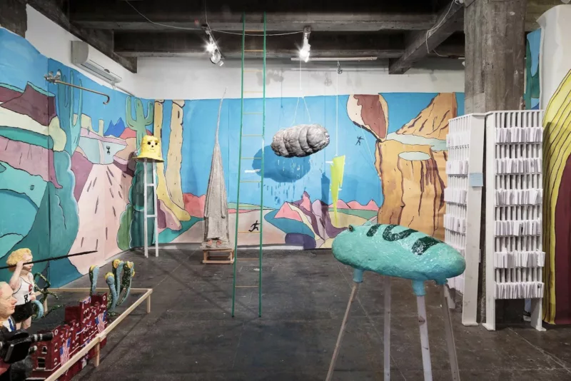 An installation shows a bright-colored abstracted landscape mural and objects scattered in a high-ceilinged space with exposed ceiling beams and a concrete floor.