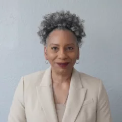 A Black woman wearing a cream colored suit jacket and complementary blouse stands in from tof a light grey wall. She is smiling broadly. She just because the Director of the Brandywine Museum and Archives