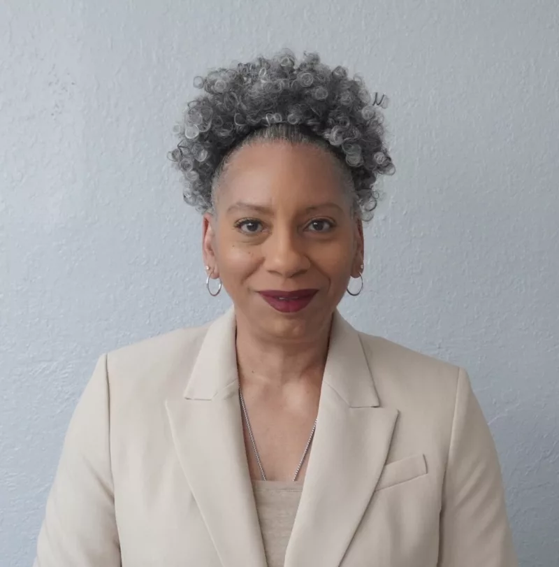 A Black woman wearing a cream colored suit jacket and complementary blouse stands in from tof a light grey wall. She is smiling broadly. She just because the Director of the Brandywine Museum and Archives