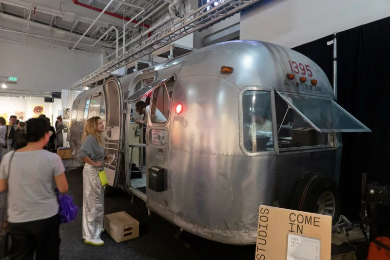 A silver Airstream trailer with its door open is parked inside the art fair. A cardboard sign announces “Worthless Studios Come In” A woman in silver mylar pants stands at the open door smiling broadly as another person passing on the left looks on, curious. Lettering on the back of the airstream says “1395 Air Stream” above a small window that is open. Several people are inside the trailer.