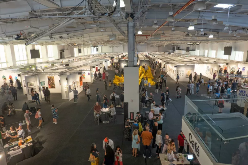 A view of a busy art fair taken from above looks down on a crowd of people walking and sitting in a large exhibition hall. A center hallway allows traffic to flow through the fairs many booths and to each side are many small booths showing art. The industrial-looking ceiling looms overhead.
