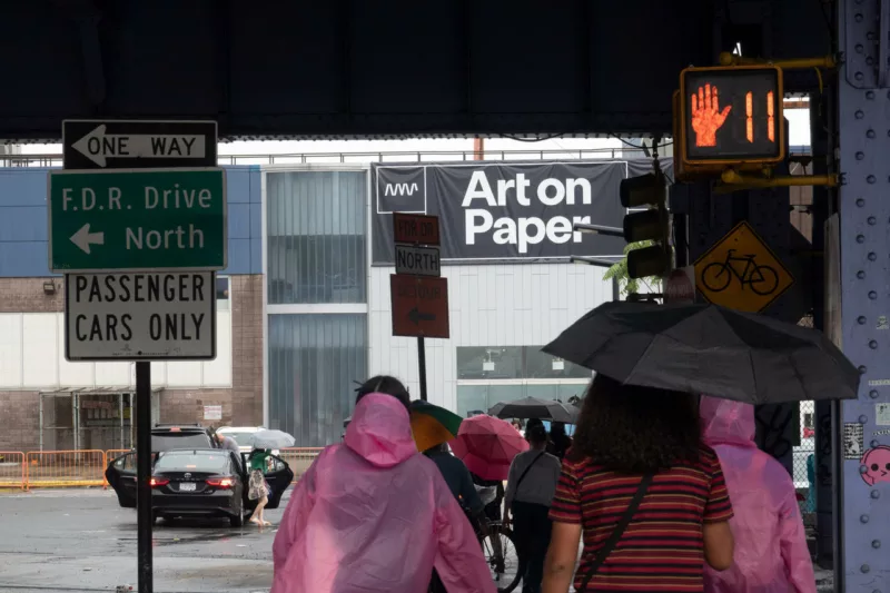 A crowd of people dressed in rain ponchos and carrying umbrellas approach Art on paper, an art fair, on a street under an underpass. Many signs feature in the photo, including a pedestrian walk sign with a hand up and 11 cautioning “don’t walk, 11 seconds left” and street directions to F.D.R Drive North, and One Way.