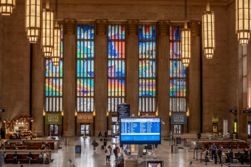A large train station waiting room in Philadelphia shows a colorful abstract design mural in the tall windows above the entrance/exit doors to the station.