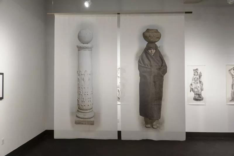 Two images side by side are printed on fabric and hanging from the ceiling. On the left is a pillar with a sphere resting on top. On the right, is an image of a woman completely swathed in dark fabric carrying a vessel or basket on her head.