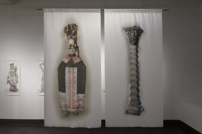 Two images side by side are printed on fabric and hanging from the ceiling. On the right is a pillar that undulates with a design that is topped with an ornamental ornament with flower and other natural motifs. On the left, is an image of a woman completely swathed in an ornamental cloth with patterns echoing the undulating pattern in the pillar. On her head is a very tall hat embellished with flowers.