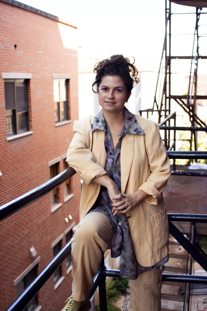 A white woman stands on a fire escape leaning on a railing, she is smiling and her dark hair is pinned up, she wears a pale peach coat.
