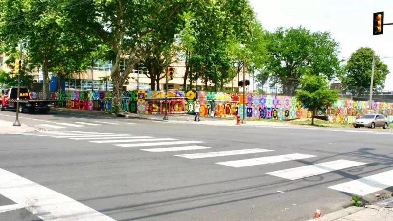 A panoramic photo shows a large urban intersection with traffic lights, people walking, cars and trucks nearby, and a sprawling wall mural that bends the corner and goes down two streets, surrounding what looks like it might be a school building or office building.