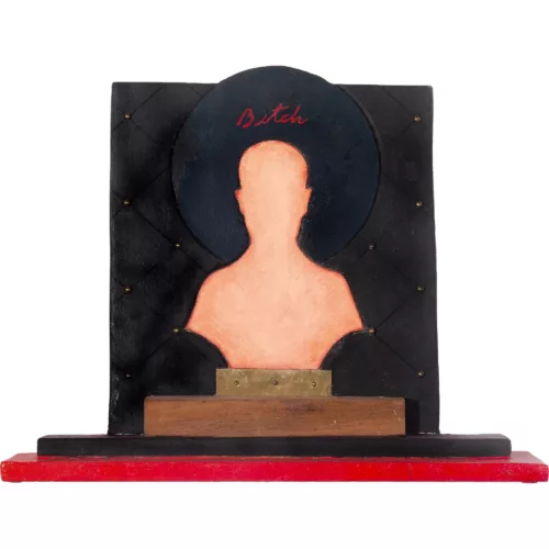 Bitch shows a pink silhouette in the center of a black background with a black halo surrounding the head and the word “Bitch” written in cursive within the halo. The piece has several blocks of wood at the base upon which the iconic work stands, like an altar.