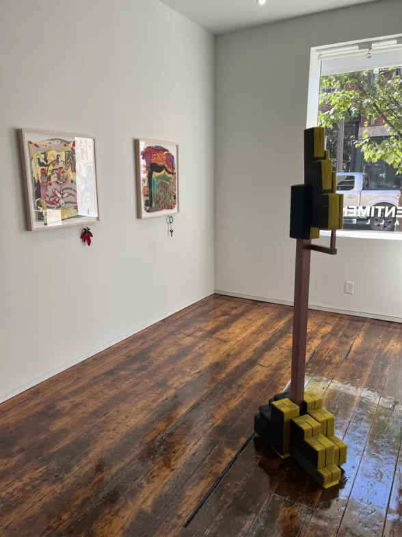A gallery with a dark wood floor and a large glass window on the street shows two works on paper framed and under glass, on the left wall. In the middle of the floor is a sculptural piece, blocky with “feet” at the bottom, a pole jutting up from the “feet” and ending in more blocks at the top and what looks like one arm bent up at the elbow giving support to the top stack of blocks
