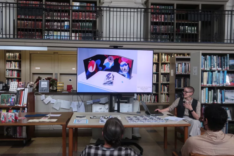 A photo shows a room in a library, with books in shelves in the background and up on a 2nd floor balcony and at a broad table covered with small books sits a young college-age white person with close-cropped hair and wearing glasses who is talking to an audience and showing slides on a screen behind them.
