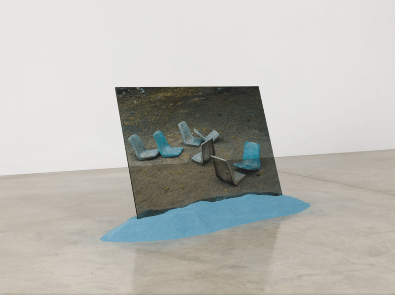 In a gallery, sitting on a polished concrete floor, is a photo printed on plexiglas that shows seven plastic, aqua blue chair tops sitting on the ground missing their legs. The photo, which is large and tilted, is embedded in a small pile of aqua-blue tinted sand. The chairs connote abandonment and the sand, perhaps, connotes a beautiful tropical island that is actually adrift and buffeted about by the ocean.
