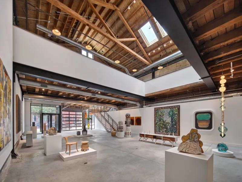 A high-ceilinged space with wood beams in a barn-like industrial building shows a new gallery space in Philadelphia, with a concrete floor and sculpture, craft objects, furniture and paintings placed in the showroom-like space. There is a skylight in the ceiling and the building’s front wall is glass, with an open, metal staircase opposite the entrance to access a balcony level above. It’s a beautiful, sophisticated and welcoming space.
