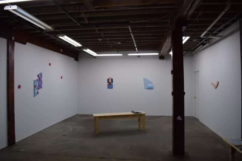 A gallery with wood beams in the ceiling and a cement floor with one wood pillar to the right shows a sparse show of art on three walls, and an austere wooden bench in the middle of the floor, with a 3 ring binder sitting on the bench.Fluorescent lights light the minimalist space.