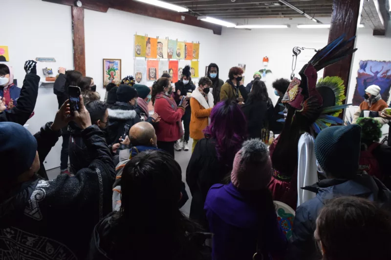 A gallery is filled with people, and with bright art works on the walls. Many people wear masks and an indigenous, costumed Aztec dancer stands to the right in the crowd. People are holding up cameras and taking pictures and videos.