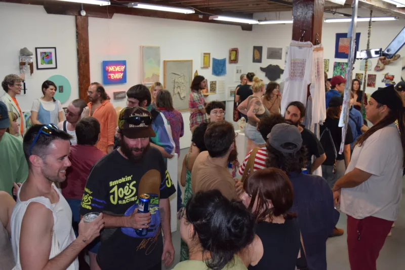 A crowd dressed for Summer in sleeveless shirts and shorts stands talking in an art gallery filled with art. They look happy and are smiling, even though it’s the last show ever at the gallery.
