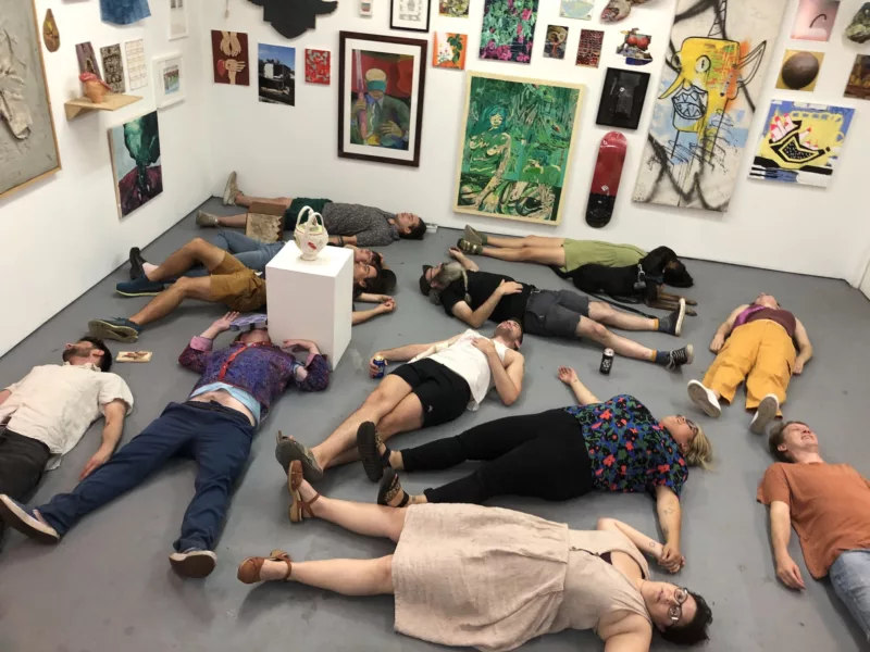 A gallery filled with art is the backdrop for a group of people who are pretending to be dead, lying down on the floor. It is 5 AM and they have been there since earlier the previous evening, celebrating and grieving over the end of the gallery.