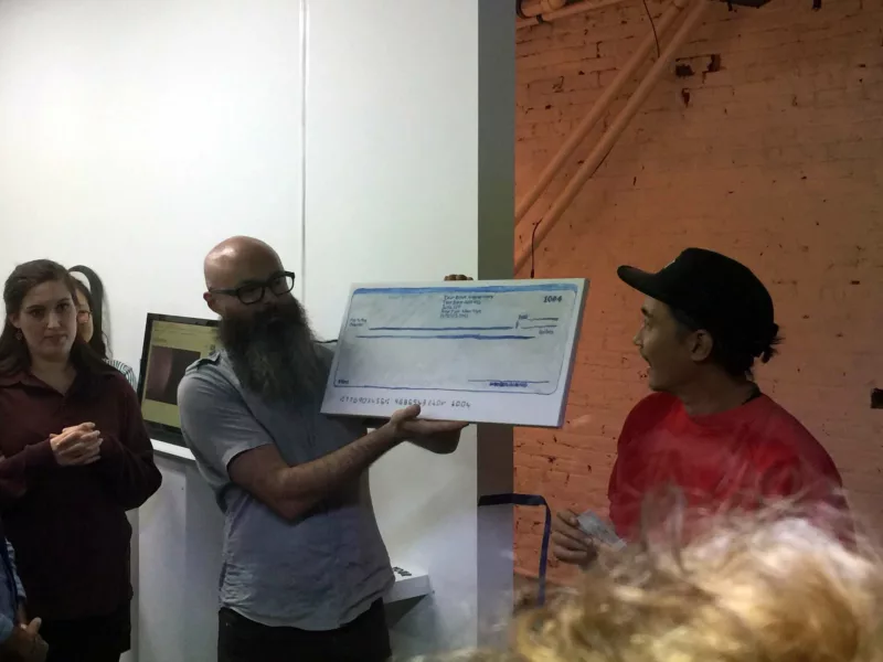 A bald-headed man with a long beard hold a poster mock up of a check for the winner of the readymade art contest at Pilot+Projects.