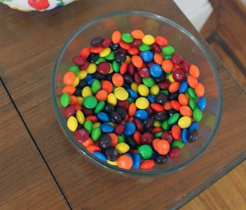 An image taken looking down at a clear glass bowl filled with Skittles and M+M candies. The bowl sits on a faux woodgrain table.