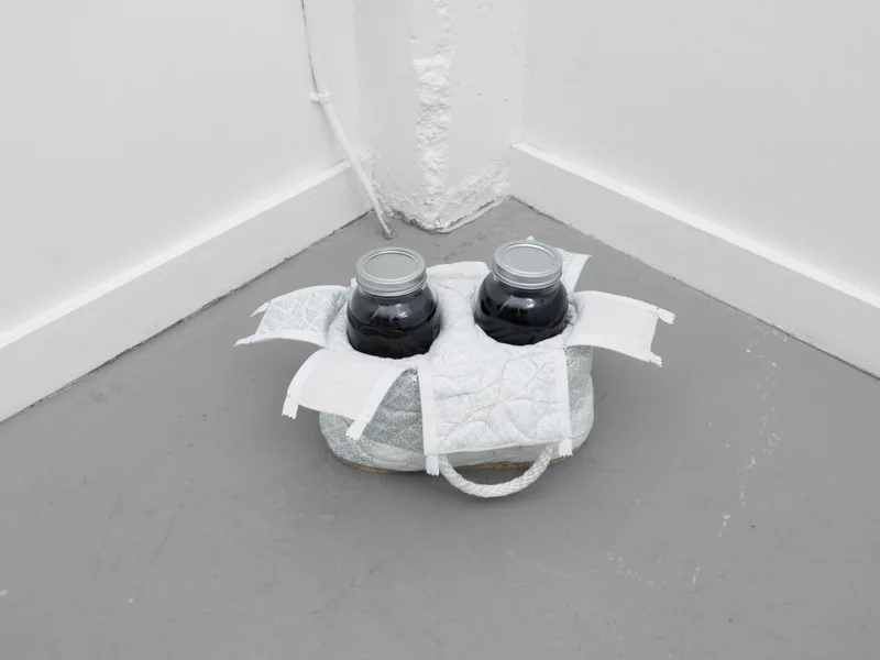 Two Mason jars, both filled with dark liquid sit on a gray-painted cement floor in a corner, in a soft-looking, quilted white carrier with rope handles. It is mysterious what they are and why they are in the corner of a room.
