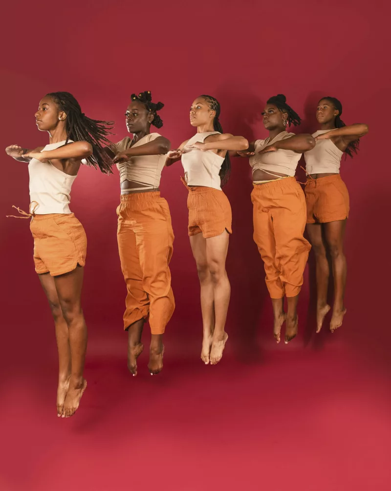 A photo shows five young Black women dancers in a row suspended in the air after having jumped in unison, their toes pointed straight down and heels in the air, their faces looking up and out and their expressions confident and determined; their orange shorts and pants and white tops complementing a solid red background of bright red. The image conveys joy, optimism and fierce determination along with beauty.