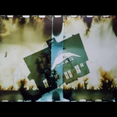 An experimental photograph shows a silhouette of a house atilt the ground with what look like burn marks or burning bushes on the ground and in the air above. A row of “white teeth” run atop and at the bottom of the picture indicating the film sprockets from analog film.