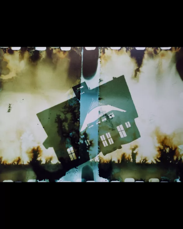 An experimental photograph shows a silhouette of a house atilt the ground with what look like burn marks or burning bushes on the ground and in the air above. A row of “white teeth” run atop and at the bottom of the picture indicating the film sprockets from analog film.