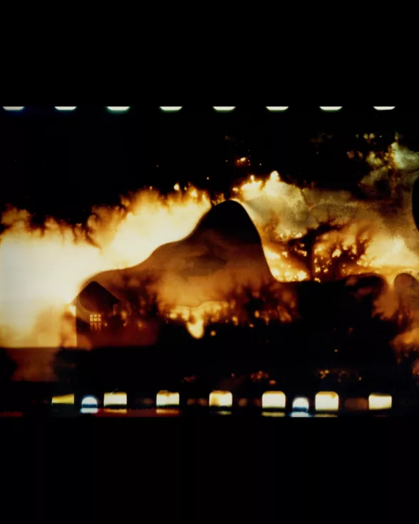 An experimental photograph shows an abstract scene, with a landscape and house suggested in the lower area and bright light, maybe from a fire, behind that and a large mountainous dark shape bracketed at the top and bottom by black space and “white teeth” indicating film sprockets from analog film. 