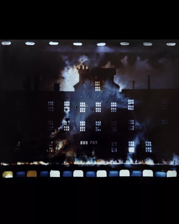 An experimental photograph shows a nighttime scene of a large institutional building, perhaps a school, in silhouette, with the windows lit up and what looks like a row of fire below and what looks like a bright area (perhaps fire) behind the silhouette. At the top, a row of “white teeth” indicate film sprockets from analog film. 