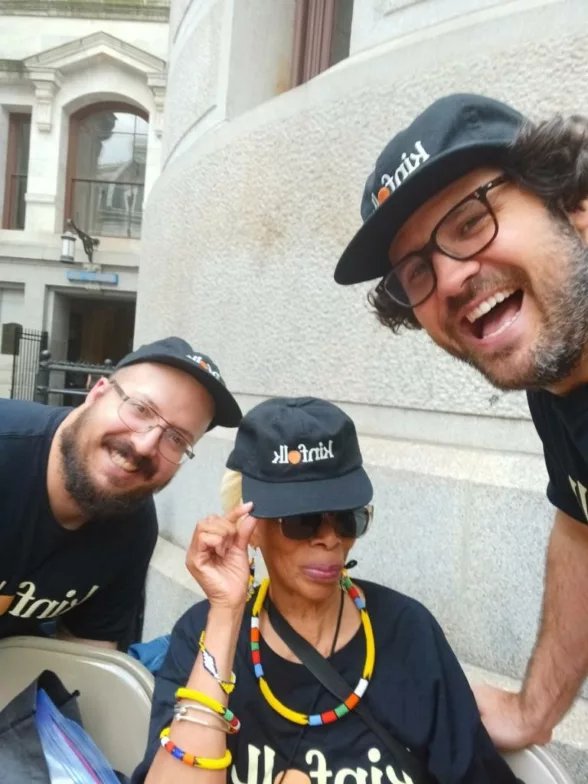 Three people pose for the camera at an event in Philadelphia. They all wear black t-shirts and hats that have a logo on them with the word “kinfolk.” The Kinfolk project is an AR monument project.