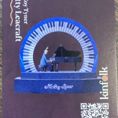 A central image shows a man in a blue suit with white hat sitting at a black grand piano where he is playing with gusto, his left hand raised in the air. The man and piano and encircled with a circlet of piano keys, and the ensemble sits atop a 2-step platform that has the name McCoy Tynor written on it. In the bottom left is a QR code to activate the AR app represented by this card. Above the QR code ar letters that spell “kinfolk,” the name of the AR monument project.