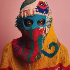 A colorful hand-knit body covering in orange, red, green, blue and pink, covers the body and head and face of an African American, whose eyes show through two holes carefully and intentionally placed in this fashionable balaclava/sweater.