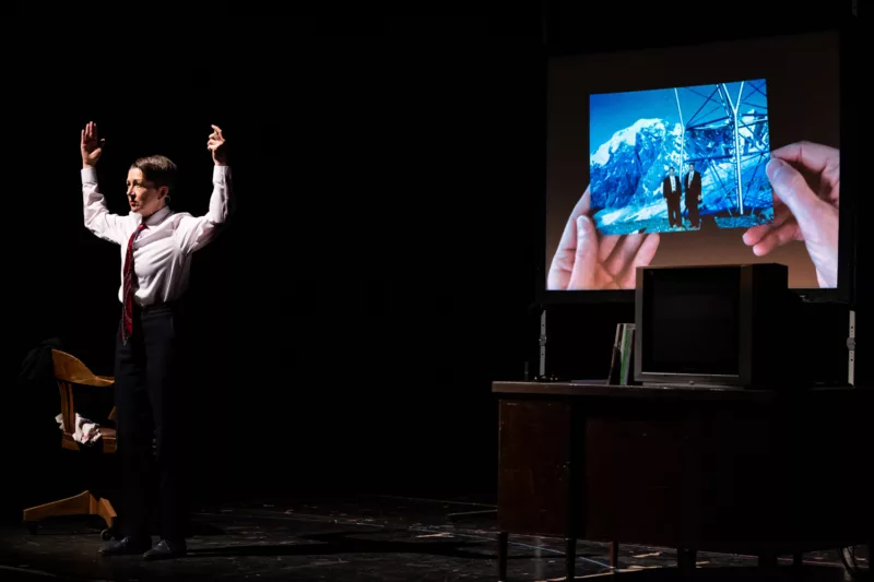 A woman stands on a stage and is brightly spot-lit. She is wearing a white shirt and a red patterned tie and her arms are raised with her elbows flexed. In back of her is a projected image of two hands holding a picture of two men in black suits standing at the base of what looks like an electricity tower in a mountainous terrain.