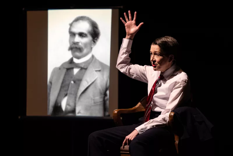 A woman sits in a chair, under a bright spotlight. She’s wearing a white shirt and a red patterned tie and is holding her right arm up, like she’s saying “Here!” Her mouth is open and she is talking. In back of her is a projection of a man in a photo from the distant past, who has a bushy mustache, and a suit coat and a vest with buttons missing.