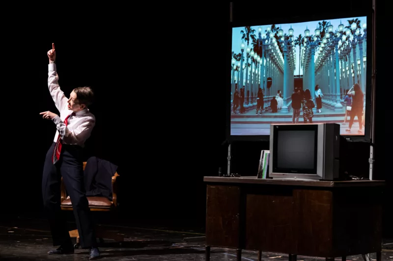 A woman stands on a stage, brightly lit, in front of a  with a desk, leather-covered chair, cathode ray tv on the desk and an image projected on the wall behind that shows a grid of 100 or more city electric light poles and people sitting and standing around them. The woman’s right arm is gesturing to the sky, her pointer finger pointing up and her left arm points forward, elbow crooked. Her mouth is open and she is talking about something related to the surrounding.