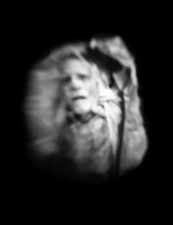 A blurry black and white photo shows a head and chest of a man who is deceased. His with eyes and mouth look open and the images is surrounded by deep black background. The image appears to be like a ghostly apparition. 