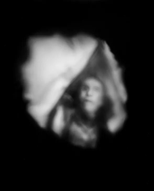 A blurry black and white photo shows a head and chest of a man who is deceased. His with eyes and mouth look open and the images is surrounded by deep black background. The image appears to be like a ghostly apparition. 